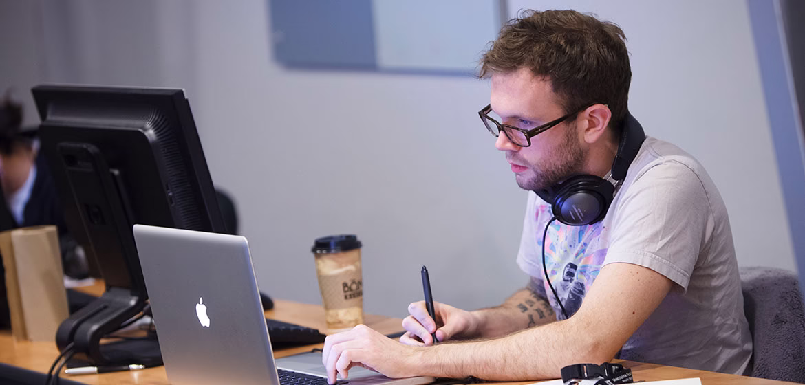 A man wearing glasses and headphones around his neck works intently on a laptop while conducting a network penetration test. He holds a stylus pen in one hand and types on the keyboard, surrounded by a desktop monitor, coffee cup, and notes, suggesting a focused cybersecurity assessment in progress.