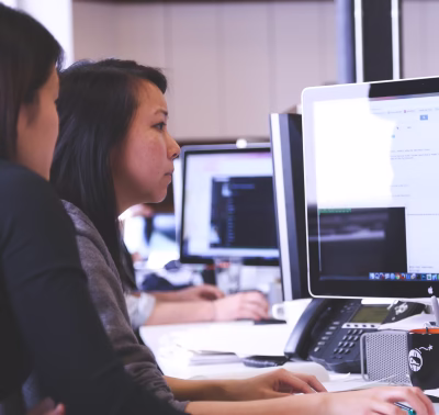 Two people sit at desktop monitors, intently reviewing code and web pages on-screen while collaborating on a web application penetration test. One person (foreground) types at the keyboard as the other looks on; visible monitors show browser windows and terminal-like code, suggesting a focused web-app security assessment in progress.